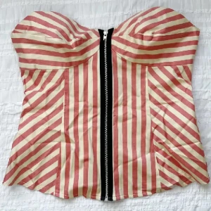 Randig korsett-topp med dragkedja - Säljer en snygg randig korsettopp i rosa och beige med en framträdande dragkedja framtill. Toppen har en stilren design med vertikala och diagonala ränder som ger en cool look. Perfekt för att lyfta vilken outfit som helst! Storlek UK 10 (XS/S).