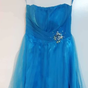 Blå långklänning med smyckesdetalj - Säljer en blå kortklänning med tunna axelband och vacker smyckesdetalj framtill i midjan. Klänningen har omlottdrapering över bysten och ett lätt, svepande tyg som ger en elegant känsla.