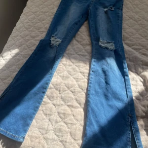 Blå bootcut jeans med slits från SHEIN - Säljer ett par blå bootcut jeans från SHEIN med slitningar på knäna och slits nertill vid bensluten. Jeansen har klassisk femficksdesign och är tillverkade i denim. Perfekta för dig som gillar en trendig och avslappnad stil.