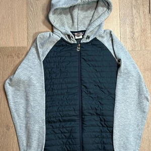 Grå och mörkblå hoodiejacka med quiltad front - Snygg hoodiejacka med grå ärmar och huva samt mörkblå quiltad front och rygg. Jackan har dragkedja hela vägen och ribbade muddar vid ärmslut och nederkant. Perfekt för lager-på-lager och en sportig look.