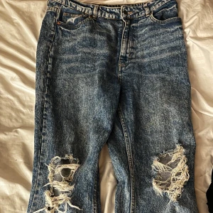 Blå slitna jeans med hål på knäna - Säljer ett par blå jeans med tydliga slitningar och stora hål på båda knäna. Jeansen har klassisk femficksdesign och normal passform. Perfekta för en avslappnad och trendig stil.