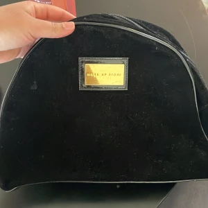 Svart necessär från Selfie Up Store - Säljer en svart necessär från Selfie Up Store. Väskan har en halvmåneform och stängs med dragkedja upptill. Perfekt för att förvara smink eller småprylar. Enkel och stilren design med en liten guldfärgad logga framtill.