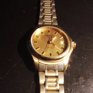 Guldfärgad klocka från Tissot - Säljer en stilren guldfärgad klocka från Tissot med metallarmband och rund urtavla. Klockan har datumvisning och klassisk design, perfekt för dig som gillar eleganta accessoarer.