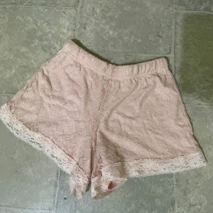 Ljusrosa spetsprydda shorts - Säljer ett par ljusrosa shorts med vacker spetsdetalj längs bensluten. Shortsen har en mjuk resår i midjan och är täckta av ett diskret blommönster i spets. Perfekta för en söt och feminin look. Perket att sova med. 
