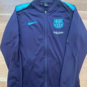 FC Barcelona träningsjacka från Nike - Säljer en mörkblå FC Barcelona träningsjacka från Nike med klubbmärke och Rakuten-logga på bröstet. Jackan har dragkedja framtill, lång ärm och turkosa detaljer på axlarna. Perfekt för fotbollsträning eller som supporterplagg.