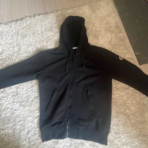 Svart hoodie från Moncler - Säljer en svart hoodie från Moncler med dragkedja och huva. Tröjan har två fickor framtill och Moncler-logga på ena ärmen. Perfekt för en stilren och avslappnad look.