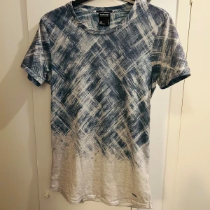 Blåvit t-shirt från Smog med abstrakt mönster - Säljer en slim fit t-shirt från Smog i storlek S. T-shirten har ett unikt blåvitt abstrakt mönster som ser ut som penseldrag och är tillverkad i mjuk bomull. Perfekt för dig som vill sticka ut med en cool och modern stil.