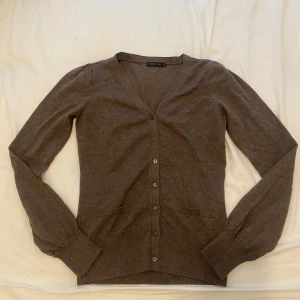 Brun kofta med v-ringning - Säljer en klassisk brun kofta från stockh lm med v-ringning och knappar framtill. Tröjan har långa ärmar och två små fickor framtill. Perfekt för lager-på-lager och enkel att matcha.