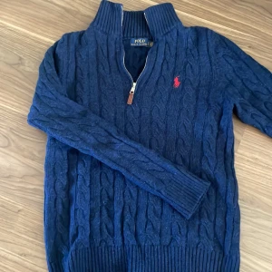 Marinblå tröja från Polo Ralph Lauren - Säljer en marinblå kabelstickad tröja från Polo Ralph Lauren med halv dragkedja och röd broderad logga på bröstet. Tröjan har hög krage och långa ärmar, perfekt för kyliga dagar. Den passar tyvärr inte mig längre. Passar storlek S 100%.