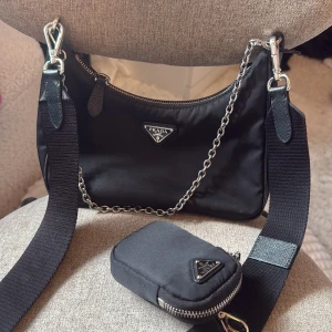 Svart axelväska från Prada med kedjedetalj - Säljer en svart axelväska från Prada med silverfärgad kedja och bred axelrem. Väskan är bara använd två gånger och har en liten avtagbar miniväska på remmen och klassisk Prada-logga framtill. Kommer med originalförpackning, dustbag och äkthetsbevis.Ny pris på väskan är 15500:-