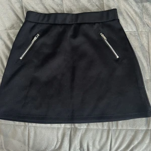 Svart kjol med dragkedjor från Lindex - Snygg svart kjol från Lindex med två silvriga dragkedjor framtill. Kjolen har en enkel, rak passform och är perfekt för en stilren look. Materialet känns mjukt och har en lätt glans.