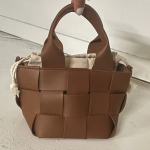 Brun flätad handväska med tygpåse - Säljer en snygg brun handväska i flätad design med korta handtag. Väskan har en innerpåse i beige tyg med dragsko. Perfekt accessoar för att lyfta din outfit! Kommer med en lång rem för att bära över dig eller på axeln 
