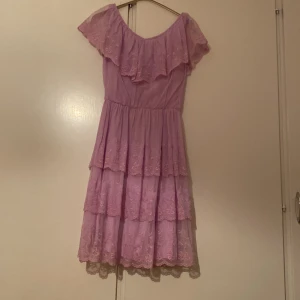 Ljuslila volangklänning med spets - Söt ljuslila klänning med volanger och spetsdetaljer. Klänningen har offshoulder-skärning och flera lager volang i kjolen. Perfekt för sommarens tillfällen och har en romantisk känsla.