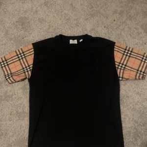 Svart t-shirt med rutiga ärmar från Burberry - Säljer en svart äkta t-shirt från Burberry med klassiska beige-rutiga ärmar. T-shirten har en normal passform och är kortärmad. Perfekt för dig som vill ha en stilren look med en touch av lyx.