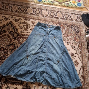 Lång jeanskjol med knappar - Säljer en blå lång jeanskjol med utsvängd passform och coola detaljer som stora fickor framtill och knappar i midjan. Kjolen har även dekorativa sömmar och spännen på sidorna för extra edge. Perfekt för dig som gillar en avslappnad men ändå trendig stil.