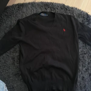 Svart v-ringad tröja från Polo Ralph Lauren - Klassisk svart v-ringad tröja från Polo Ralph Lauren i mjuk bomull. Tröjan har lång ärm och en liten röd broderad logga på bröstet. Perfekt för en stilren och enkel look. 