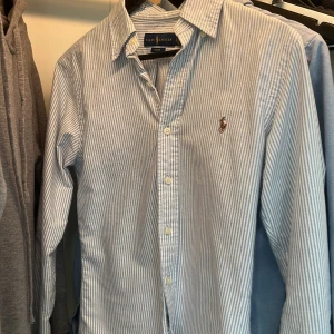  Ralph Lauren  - Säljer en klassisk blårandig skjorta från Ralph Lauren i slim fit-modell. Skjortan har långa ärmar, knappar framtill och den ikoniska broderade loggan på bröstet. Perfekt för en stilren och tidlös look. Storlek smal