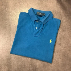 Blå piké från Polo Ralph Lauren - Säljer en klassisk blå pikétröja från Polo Ralph Lauren med gul broderad logga på bröstet. Tröjan har krage, knappar framtill och är kortärmad. Perfekt för en stilren och avslappnad look. Tröjan är i storlek M och har lite smuts som ni ser på bild 5. Bara att höra av sig vid funderingar eller frågor😀