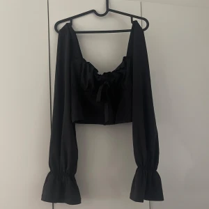 Svart croppad topp med knyt och långa ärmar - Söt långärmad top med fina detaljer, perfekt till sommaren. Rosett/knytdetalj framtill. Dragkedja bak. Storlek L (40/42), låg fyrkantig urringning. Helt ny och oanvänd.