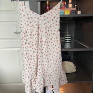 Vit blommig kortklänning med volang - Supersöt vit kortklänning med små rosa blommor och volang nertill. Klänningen har smala axelband och v-ringning framtill. Perfekt för varma dagar och har en luftig, lätt känsla. Använd en gång❤️