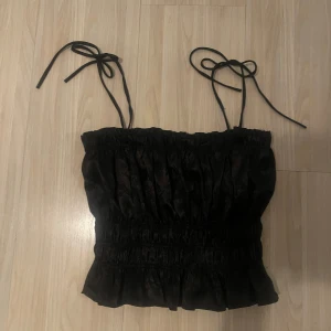 Svart croppad linnetopp med knytband - Säljer en svart croppad linnetopp med smala axelband som knyts upptill. Toppen har smockad midja och volangkant nedtill, vilket ger en söt och trendig look. Perfekt att styla till jeans eller kjol.