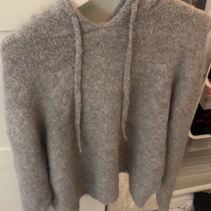 Grå stickad hoodie - Mysig grå stickad hoodie med luva och dragsko. Tröjan har lång ärm och är tillverkad i ett mjukt material som passar perfekt för kyligare dagar.