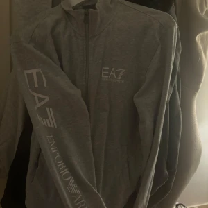 Grå ziptröja från emporio Armani -  grå ziptröja från Emporio Armani  med logga på bröstet och tryck längs ena ärmen. Tröjan har krage, dragkedja framtill och två fickor. Storlek M