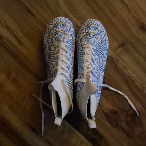 Blå och vita fotbollsskor med mönster - Säljer ett par fotbollsskor med högt skaft i blått, vitt och gult mönster. Skorna har snörning och en riktigt cool sula i guld och rött. Perfekta för dig som vill sticka ut på planen! Knappt använda, köpte för cirka 200kr