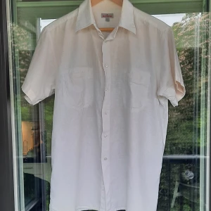 Vit kortärmad Batistini skjorta - Säljer en klassisk vit skjorta från Batistini i storlek L. Skjortan är kortärmad med två bröstfickor och knäppning framtill. Tillverkad i en luftig mix av linne och bomull, perfekt för varma dagar. Pris kan diskuteras