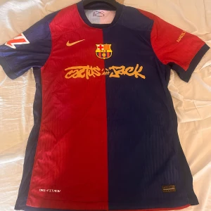 FC Barcelona x Nike fotbollströja Lamine Yamal 19 - Säljer en FC Barcelona fotbollströja i rött och blått med gult tryck, Nike-logga och klubbmärke. Tröjan har korta ärmar och nummer 19 Lamine Yamal på ryggen. Unik design med Cactus Jack-tryck framtill.