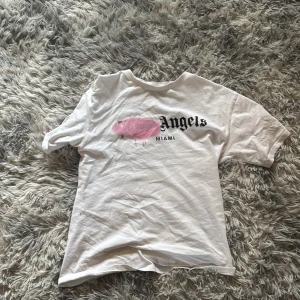 Vit t-shirt från Palm Angels med tryck - Vit t-shirt från Palm Angels med svart text 'Angels' och 'MIAMI' på bröstet. Det finns även en rosa spray paint-effekt över loggan. Klassisk passform och rund halsringning. Perfekt för en streetwear-look.