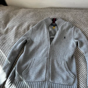 Grå stickad kofta från Polo Ralph Lauren - Säljer en klassisk grå stickad kofta från Polo Ralph Lauren med dragkedja framtill och två fickor. Tröjan har ribbade muddar och en liten broderad logga på bröstet. Perfekt för lager-på-lager-stil.