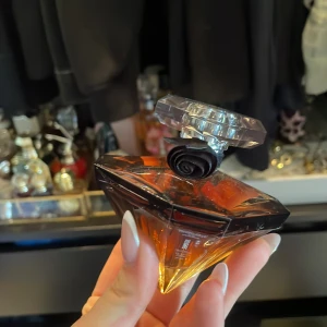 La Nuit Trésor Eau de Parfum - En lyxig parfym från Lancôme i en diamantformad flaska med svart rosdetalj vid halsen. Flaskan har en gradient från mörkbrun till bärnstensfärgad och ett transparent lock. Doften är sensuell och elegant.