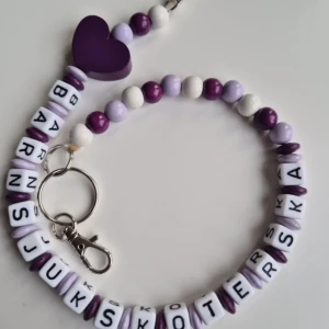 Lila och vit nyckelkedja med hjärta och bokstavspärlor - Nyckelkedja med pärlor i lila, vitt och mörklila samt ett stort lila hjärta. Bokstavspärlor bildar ordet 'Barnsjuksköterska'. 