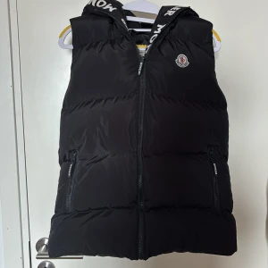Svart dunväst från Moncler med huva - Svart dunväst från Moncler med huva och dragkedja framtill. Västen har två fickor med dragkedja och Moncler-logga på bröstet. Insidan av huvan har Moncler-tryck. Perfekt för lager-på-lager under kyliga dagar.