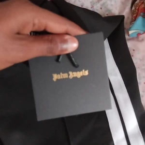 Svart träningsjacka från Palm Angels - Säljer en svart träningsjacka från Palm Angels med vita ränder längs ärmarna och logga på bröstet. Jackan har dragkedja och sportig look, perfekt för dig som gillar streetwear.