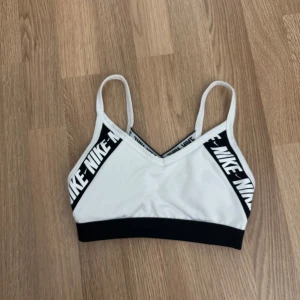 Vit och svart sport-bh från Nike - Sport-bh från Nike i vitt och svart med tunna axelband och bred resår under bysten. Nike-logga i vitt på svarta band längs kanterna. Perfekt för träning och har en stilren design. storlek XS. 50kr
