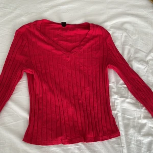 Ribbad röd långärmad tröja med v-ringning - Säljer en snygg röd långärmad tröja med v-ringning och ribbad struktur. Perfekt att styla till jeans eller kjol för en trendig look. Mjuk och stretchig i materialet för en bekväm passform. Storleken är L men skulle kunna även passa M eller S.