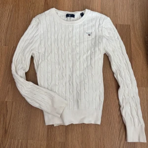 Vit kabelstickad tröja Gant - Säljer en klassisk vit stickad tröja från Gant med rund halsringning och diskret logga på bröstet. Tröjan har ett kabelstickat mönster och långa ärmar. Perfekt för en stilren och tidlös look. Storlek 158/164. 70kr