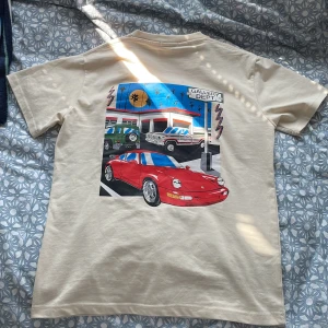 Gallery Dept T-shirt med Porschetryck - Vit/Cream/Beige/Offwhite t-shirt från Gallery Dept. Med färgglatt tryck på ryggen som visar en röd Porsche och bensinstation. Liten logga och adress på bröstet. Klassisk passform och rund halsringning. Storlek M men passar S. Använd en gång och säljer för att den var för stor.
