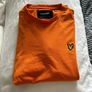 Orange t-shirt från Lyle & Scott - Snygg orange t-shirt från Lyle & Scott med klassisk rund hals och broderad gul logga på bröstet. 
