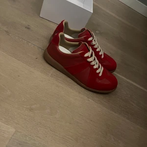 Röda sneakers från Maison Margiela - Säljer ett par röda sneakers från Maison Margiela med beige snörning och gummisula. Skorna har en klassisk rund tå och är tillverkade i en mix av mocka och skinn. Perfekta för dig som vill sticka ut med en färgstark look.
