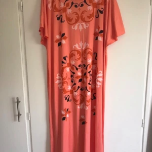 Kaftan  - Säljer en korallfärgad koftan med korta ärmar och dekorativt mönster i vitt, svart och orange. Klänningen har en rak och lös passform, perfekt för varma dagar eller avslappnade tillfällen.