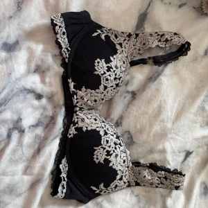 Svart intimissimi bh - Snygg svart bh med vit spets och blommigt mönster. Axelbanden är dekorerade med spets och bh:n har hakknäppning bak. Perfekt för dig som vill ha något extra fint under kläderna.