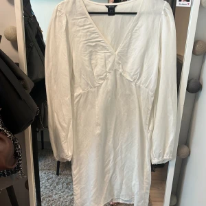 Vit långärmad klänning från Lindex - Säljer en stilren vit klänning från Lindex med lång ärm och v-ringad hals. Klänningen har en mjuk och lätt glansig yta, vilket ger en elegant känsla. Perfekt för dig som gillar enkel och klassisk stil.