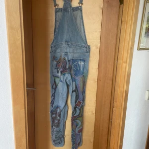 Jeanshängselbyxor med graffiti-mönster - Unika blå jeanshängselbyxor med färgglada graffiti-inspirerade målningar och slitna detaljer på benen. Byxorna har klassisk hängselmodell med fickor fram och bak, samt raka ben. Perfekt för dig som vill sticka ut med din stil.