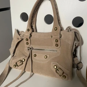 Beige axelväska i mocka med gulddetaljer - Säljer en beige axelväska i mocka som är lik balenciagas väskor med guldfärgade nitar och dragkedja framtill. Väskan har både handtag och axelrem samt en liten spegel som accessoar. Perfekt för dig som vill ha en snygg och praktisk väska.