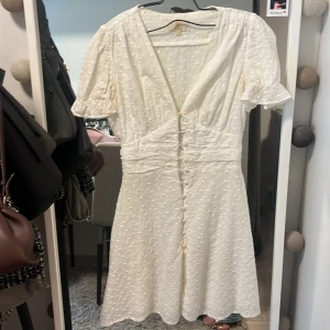 Vit kortklänning med blommigt mönster från Chelsea - Supersöt vit kortklänning från Chelsea med små broderade blommor, v-ringning och knappar framtill. Puffiga kortärmar och markerad midja ger en romantisk känsla. Perfekt för sommarens alla tillfällen!