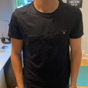 Svart t-shirt från GANT - Säljer en klassisk svart t-shirt från GANT i storlek M. T-shirten har rund halsringning och liten broderad GANT-logga på bröstet. Perfekt basplagg till garderoben. Säljer eftersom att de har blivit för liten.