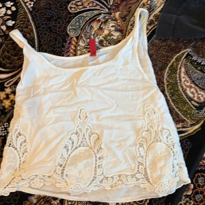 Vit linnetopp med spets från H&M Divided - Säljer en vit linnetopp från H&M Divided med breda axelband och dekorativa spetsdetaljer längs nederkanten. Perfekt för sommardagar och enkel att matcha med jeans eller kjol.